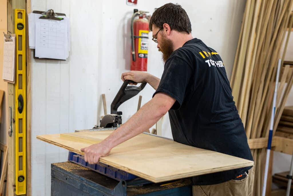 TressCo Construction team member with glasses, beard, and back TressCo Construction t-shirt, measuring a piece of wood to be used in custom cabinetry.