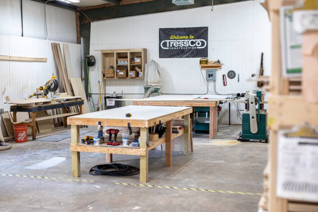 Shot of the work area at TressCo Construction's Cabinet Shop. There are Tables to work at, a saw, and various other tools and materials. Also features a sign that says, "Welcome to TressCo Construction."